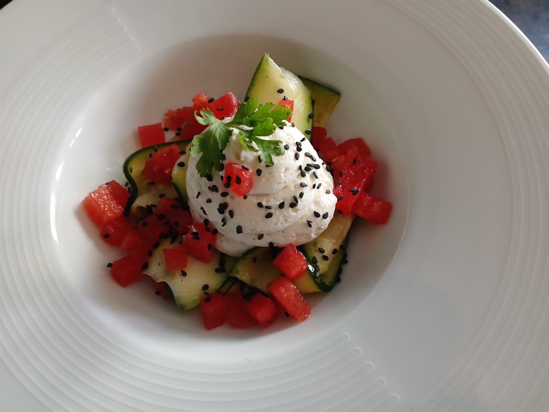 Salade de pastèque et courgettes marinées, espuma de fromage frais au sésame noir, dans une assiette creuse blanche