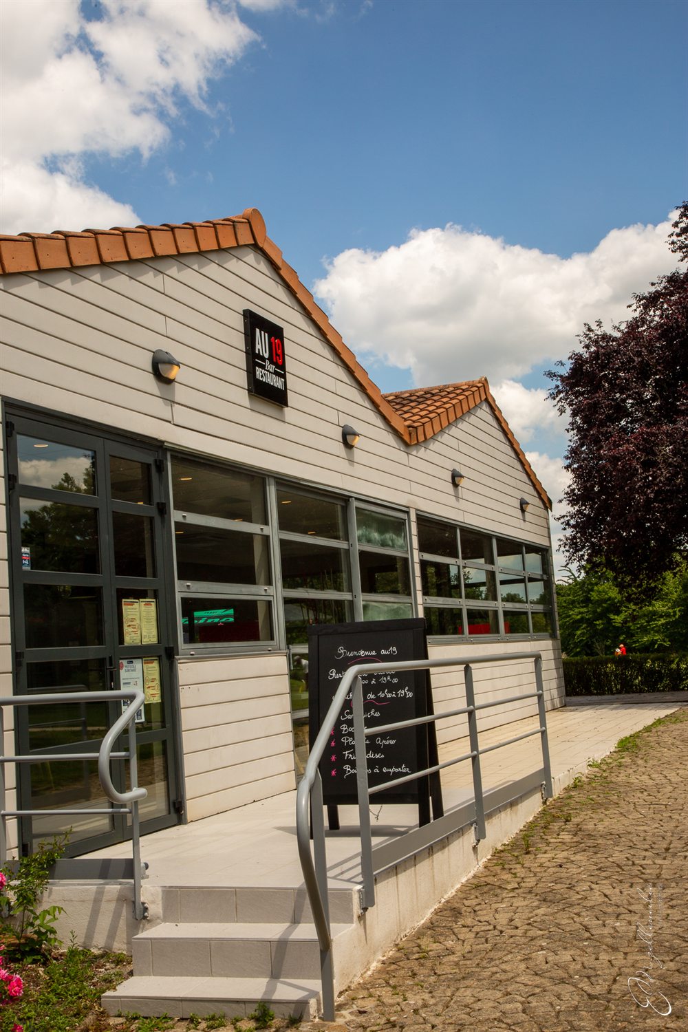L’entrée du restaurant, sa rampe d’accès et l’ardoise du jour, sous un ciel bleu